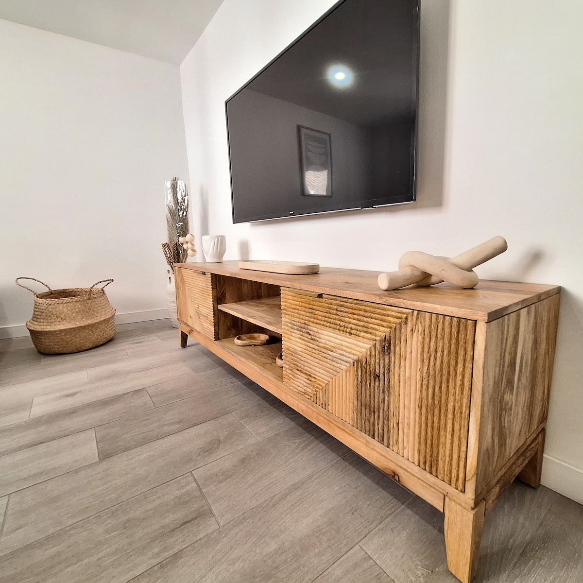 A wooden TV cabinet stands against a white wall, featuring a smooth surface and geometric patterns. A flat-screen television is mounted above, providing a focal point in the room. A woven basket sits nearby, adding texture to the space.