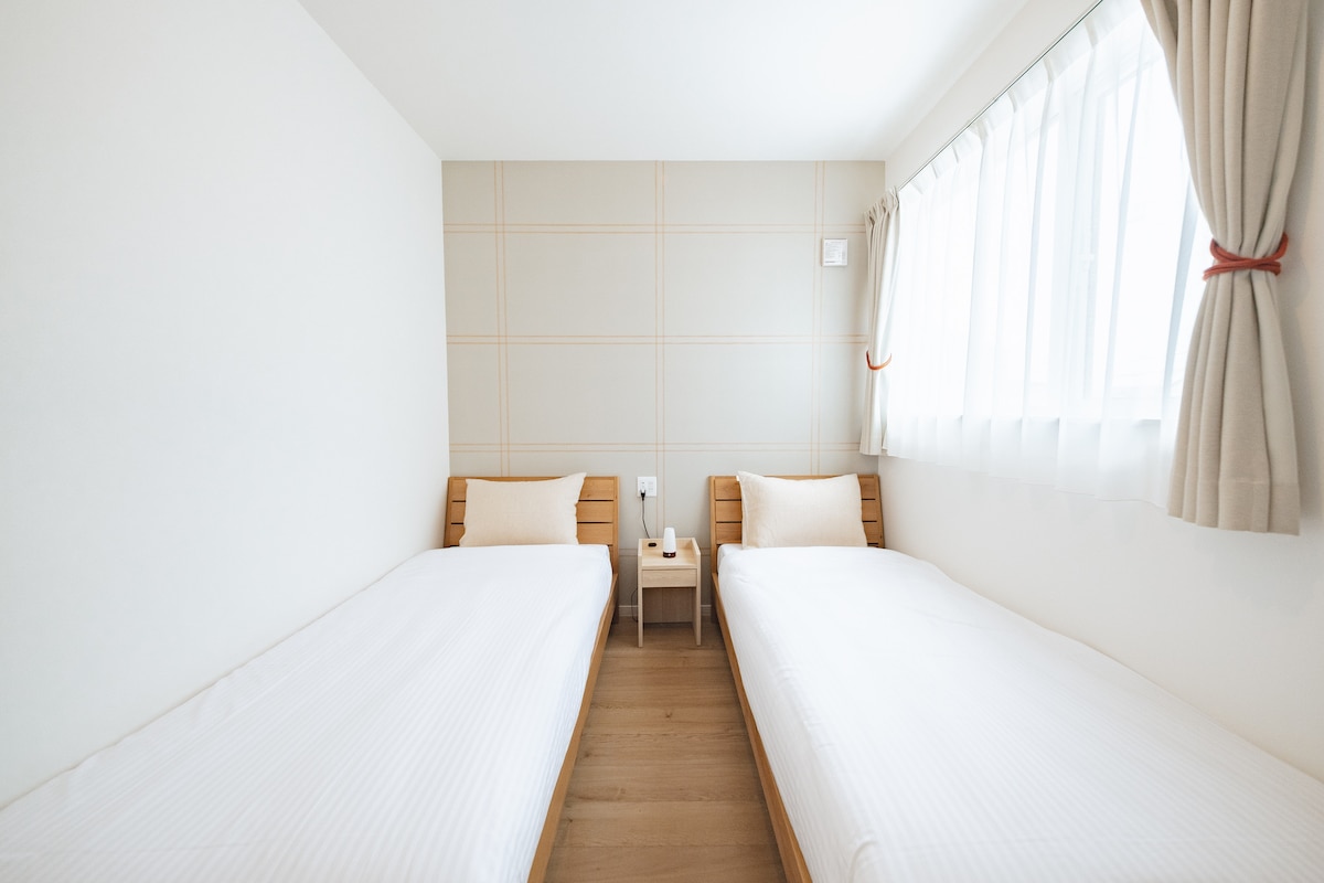 The image depicts a simple and functional guest room with two twin beds arranged parallel to each other. A small wooden table sits between the beds, with a lamp placed on top. Natural light filters through a window dressed with light-colored curtains.