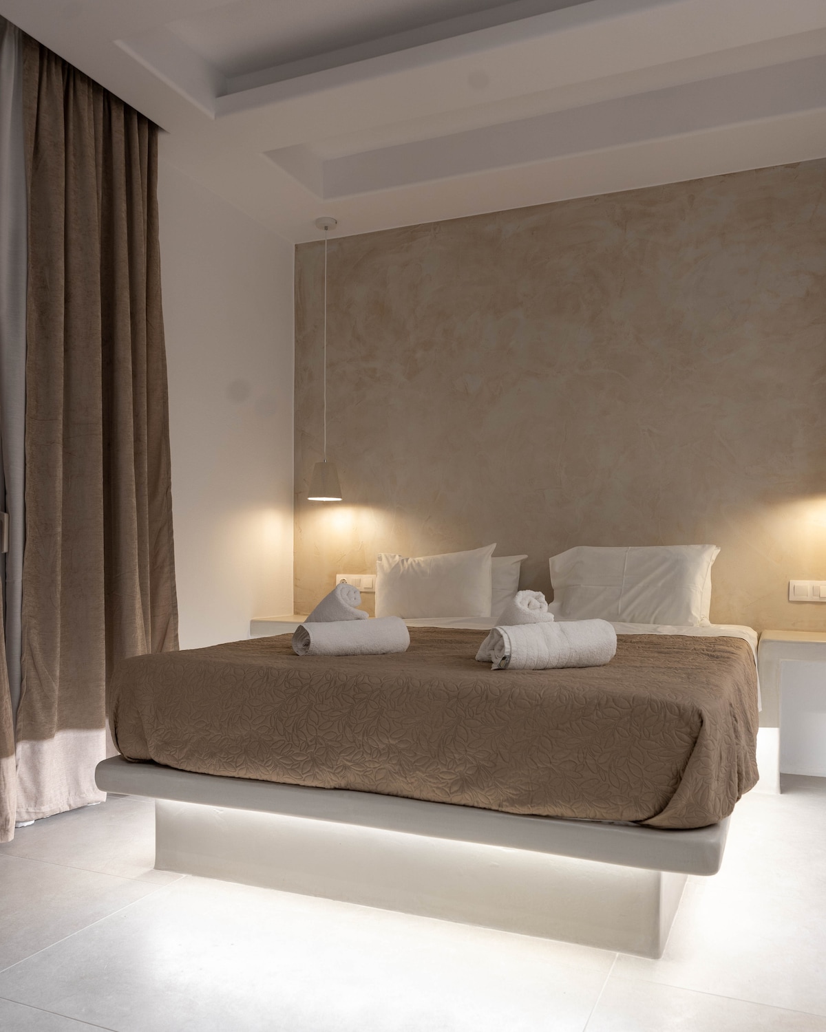 A cozy sleeping area features a spacious bed with a textured brown coverlet and white pillows. Soft lighting from two hanging fixtures provides a gentle glow. The walls present a smooth, light-colored finish, and natural light filters through a large curtain draped beside the bed.