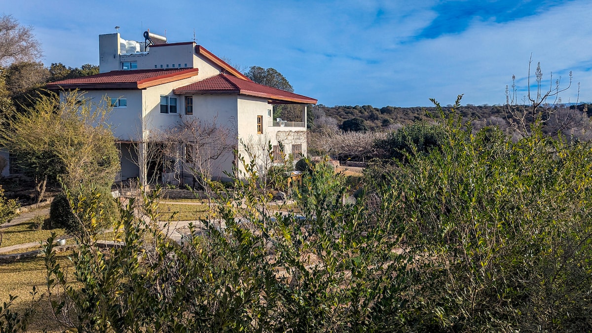 A two-story house is surrounded by lush greenery, featuring a red-tiled roof and light exterior walls. The structure is positioned within a spacious landscape that includes well-maintained gardens and pathways, offering a serene environment.