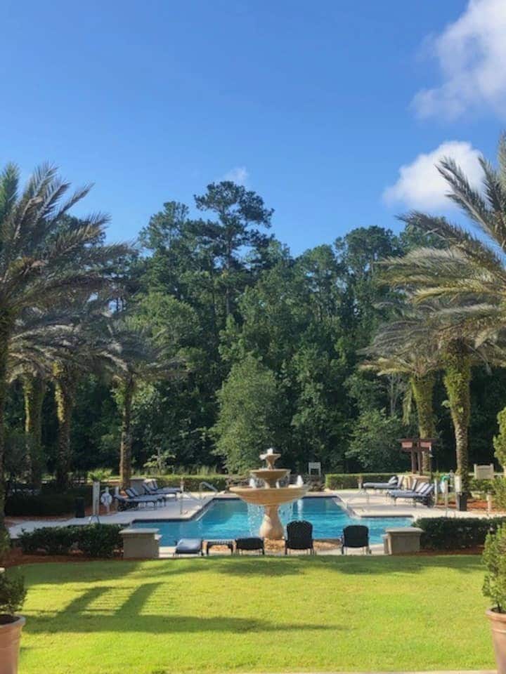 A sparkling pool is surrounded by lush greenery and palm trees, providing a serene outdoor space. A central fountain adds a touch of elegance. Lounge chairs are placed around the pool, inviting relaxation in a tranquil setting.