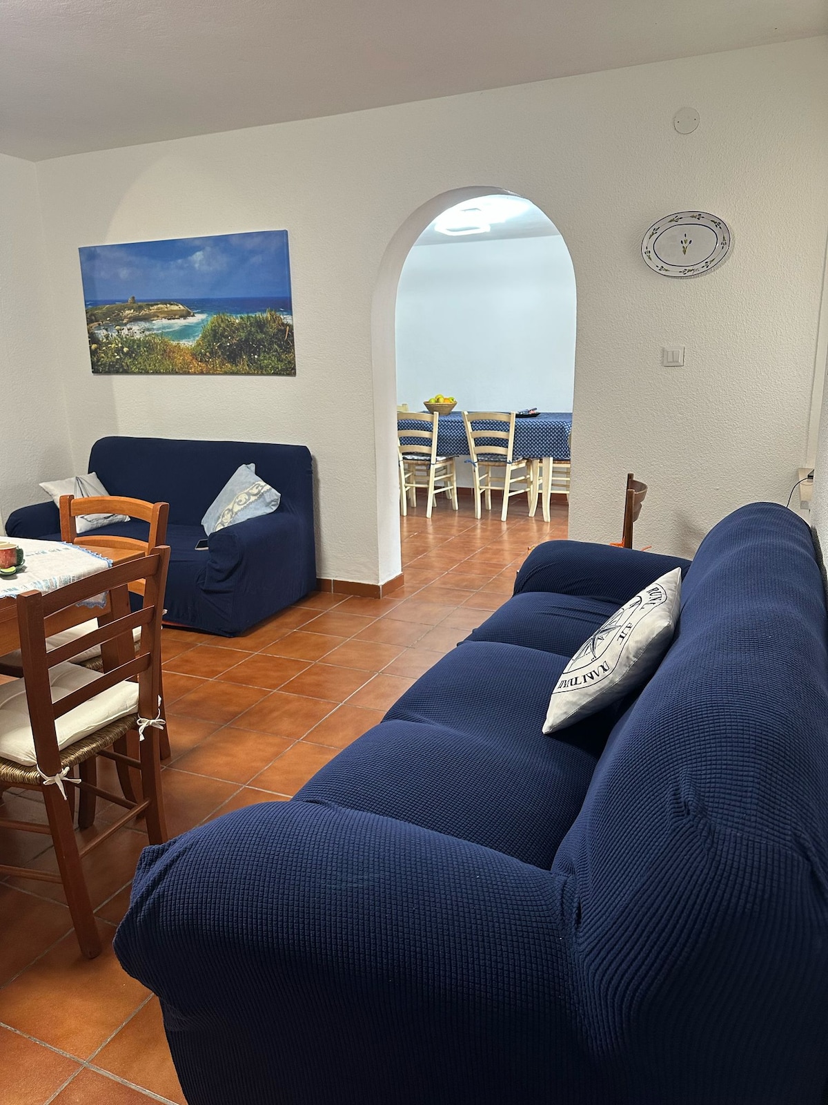 The living area showcases a navy blue sofa and a wooden dining table with light chairs. A colorful artwork depicting a coastal scene hangs on the wall, while an arched entry leads to a dining space set with a blue tablecloth and fresh flowers.