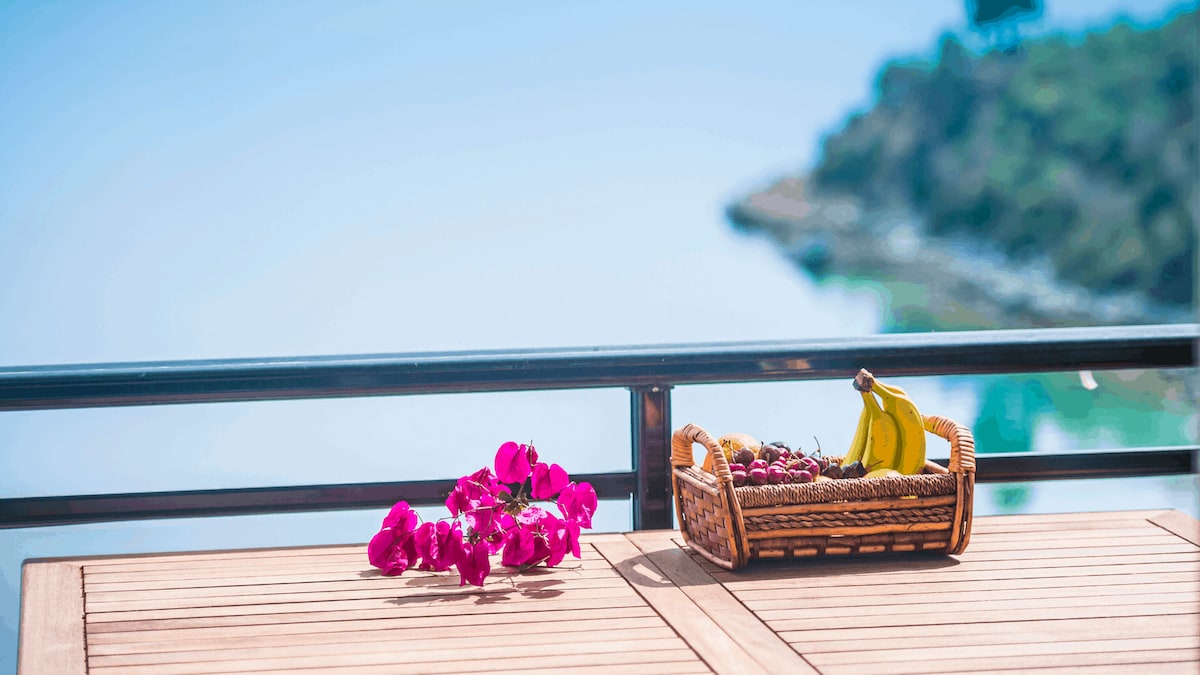 A wooden table is adorned with a woven basket of fruit, including bananas and grapes. Pink bougainvillea blossoms are placed nearby, while the background showcases a serene sea view and lush greenery along the coastline.