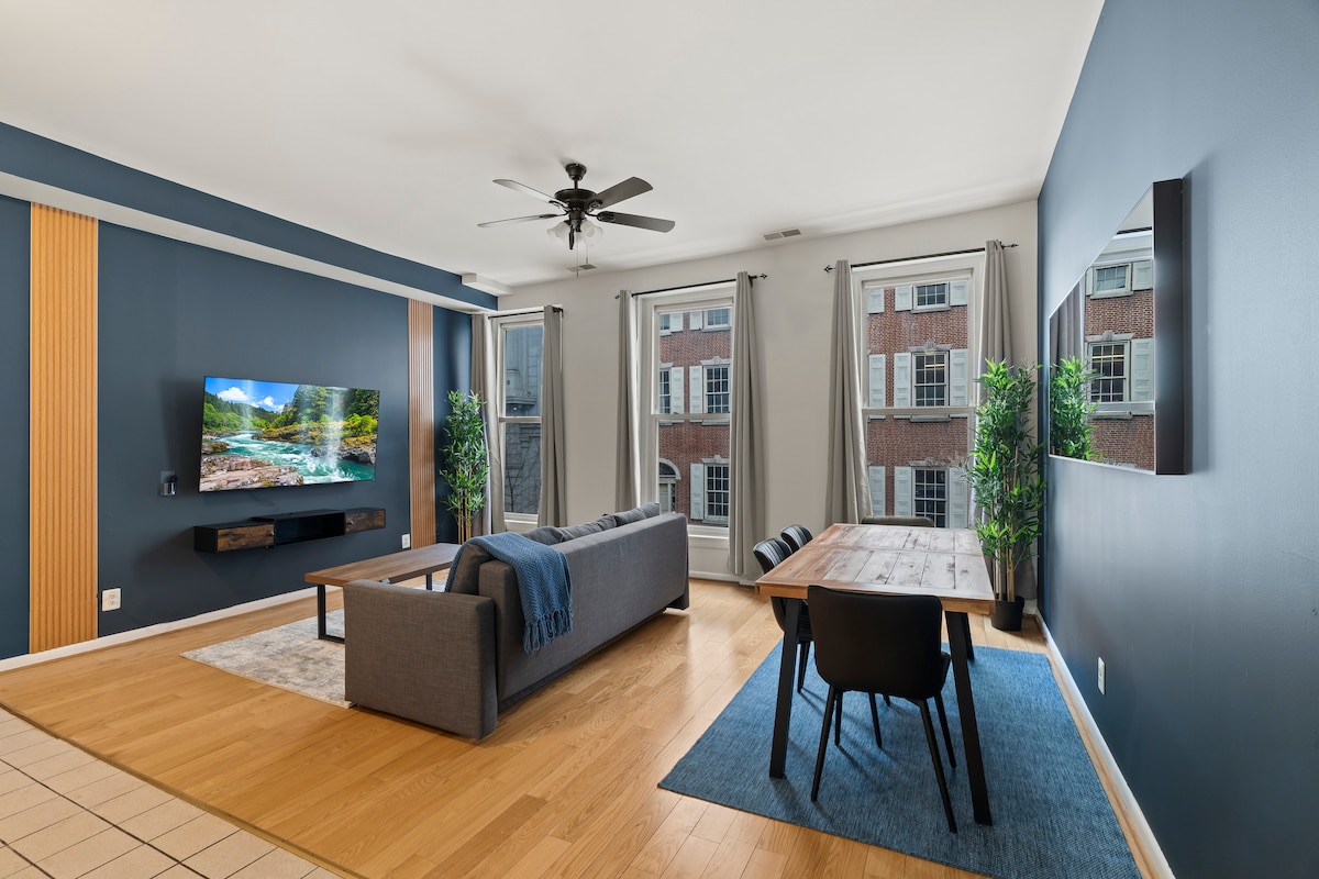 A living area features a comfortable couch positioned beside a wood dining table with chairs. A large wall-mounted TV displays a scenic image. Natural light enters through two windows, framed by light curtains, illuminating the light wood flooring.