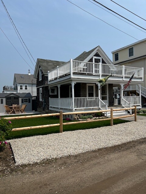 The exterior of a two-story beach house is displayed, featuring a classic coastal design with stone and wood elements. Two wraparound decks provide outdoor space for seating. A gravel driveway leads to a well-maintained front yard with decorative landscaping and an American flag hanging by the entrance.