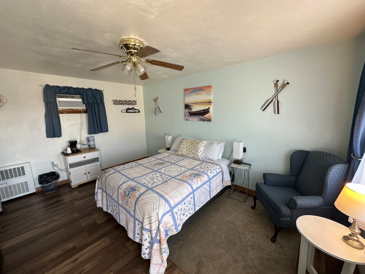 A guest room features a spacious bed with a patterned quilt, complemented by a blue armchair. Wall decorations include a serene landscape print and two decorative items. Soft lighting comes from two bedside lamps, with a window dressed in blue curtains allowing natural light.