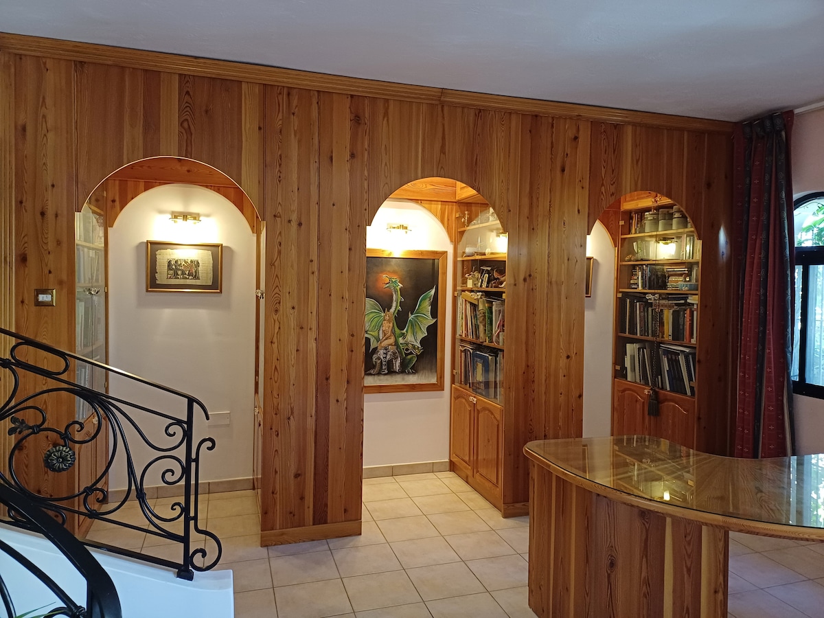 Wooden paneling surrounds an inviting nook featuring arched openings that lead to a collection of bookshelves. A colorful painting depicting a dragon is central, framed by warm lighting from wall sconces. The tiled floor complements the overall design, ensuring a cozy atmosphere.