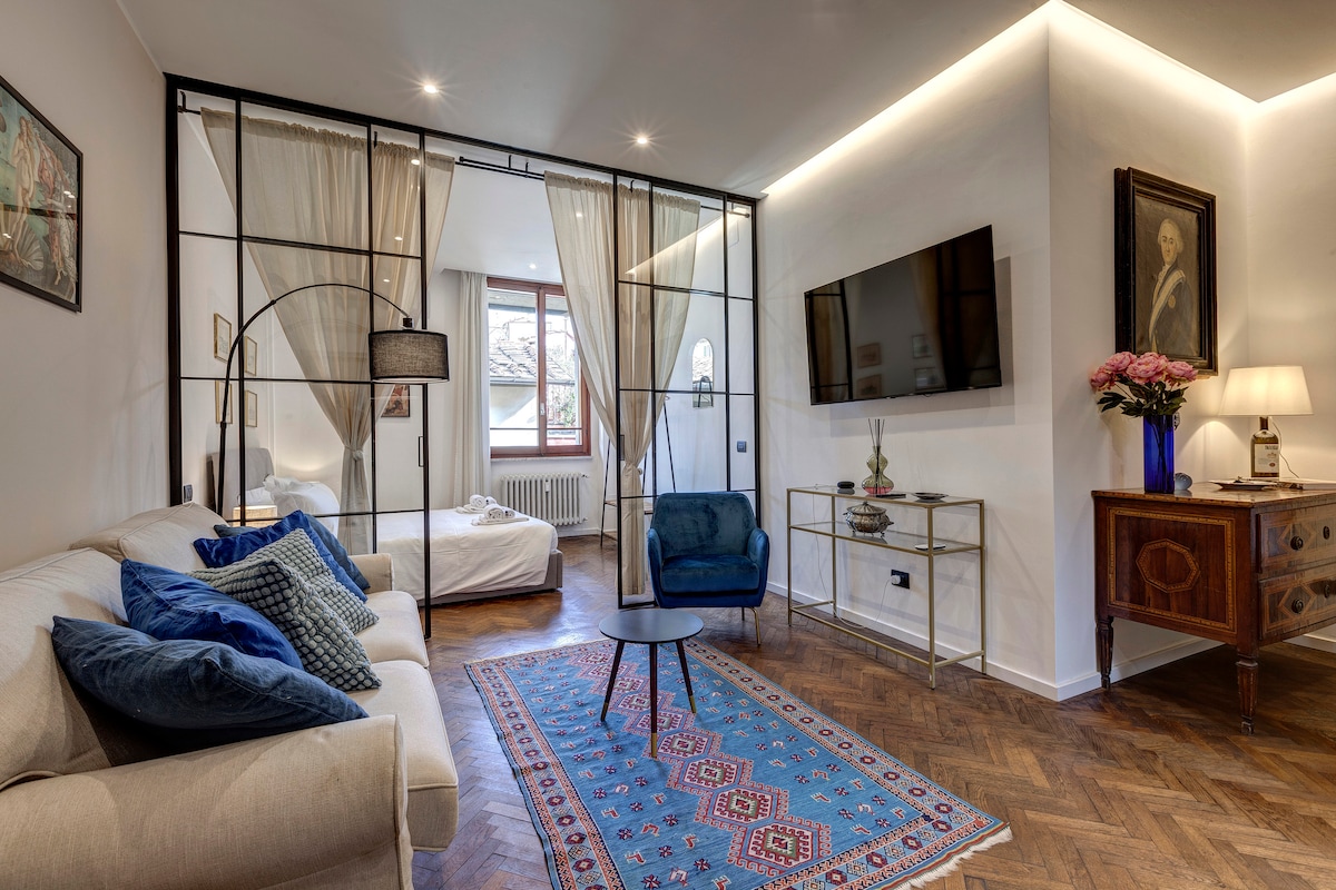 A stylish living area features a soft sofa adorned with blue cushions, providing a cozy space. A modern blue chair and a small table complement the decor. Elegant glass partitions separate the living space from the bedroom, which is illuminated by natural light from the window.