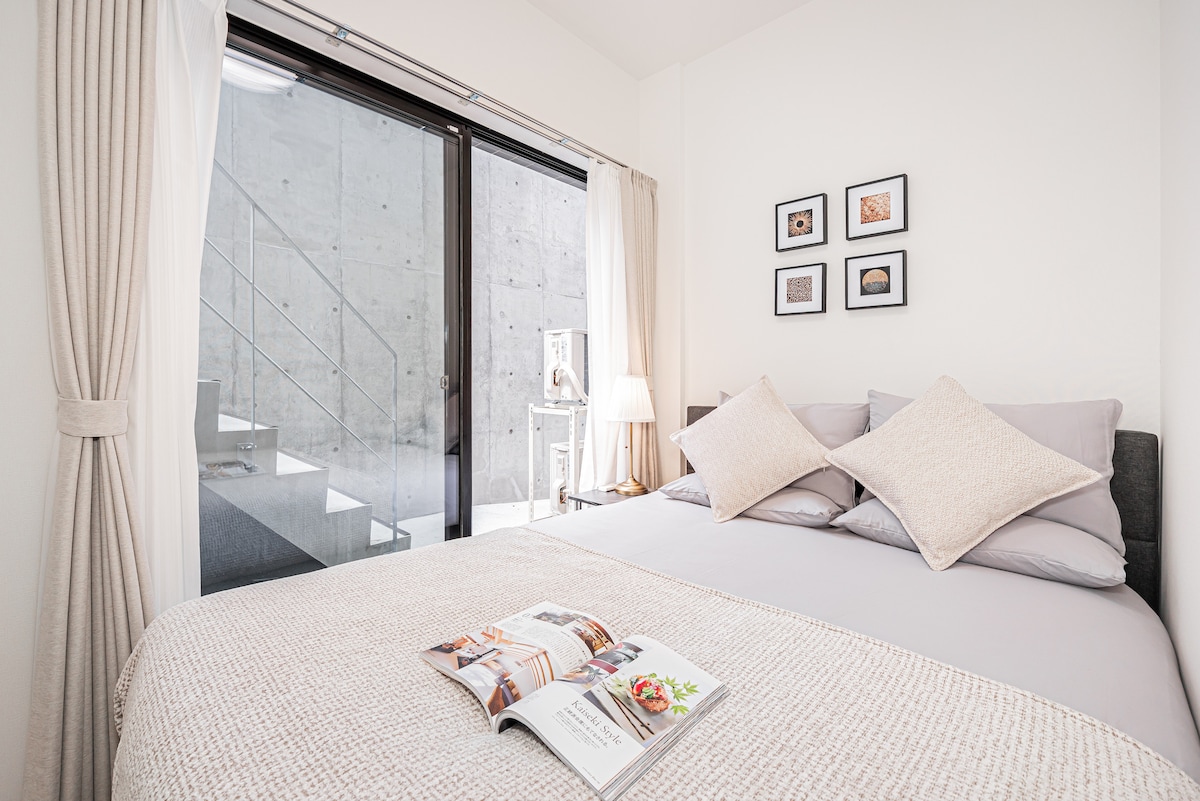 A neatly arranged bedroom features a spacious bed with soft gray linens and decorative pillows. Natural light filters in through large windows framed by soft curtains, illuminating the minimalist decor, including framed artwork on the wall. A magazine lies open on the bed.