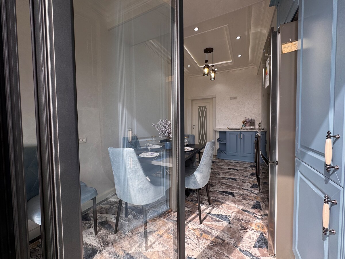 A bright dining area is visible through expansive glass doors. Light blue chairs surround a dark wooden table, set for a meal. Elegant pendant lighting hangs above, while textured flooring complements the overall decor.