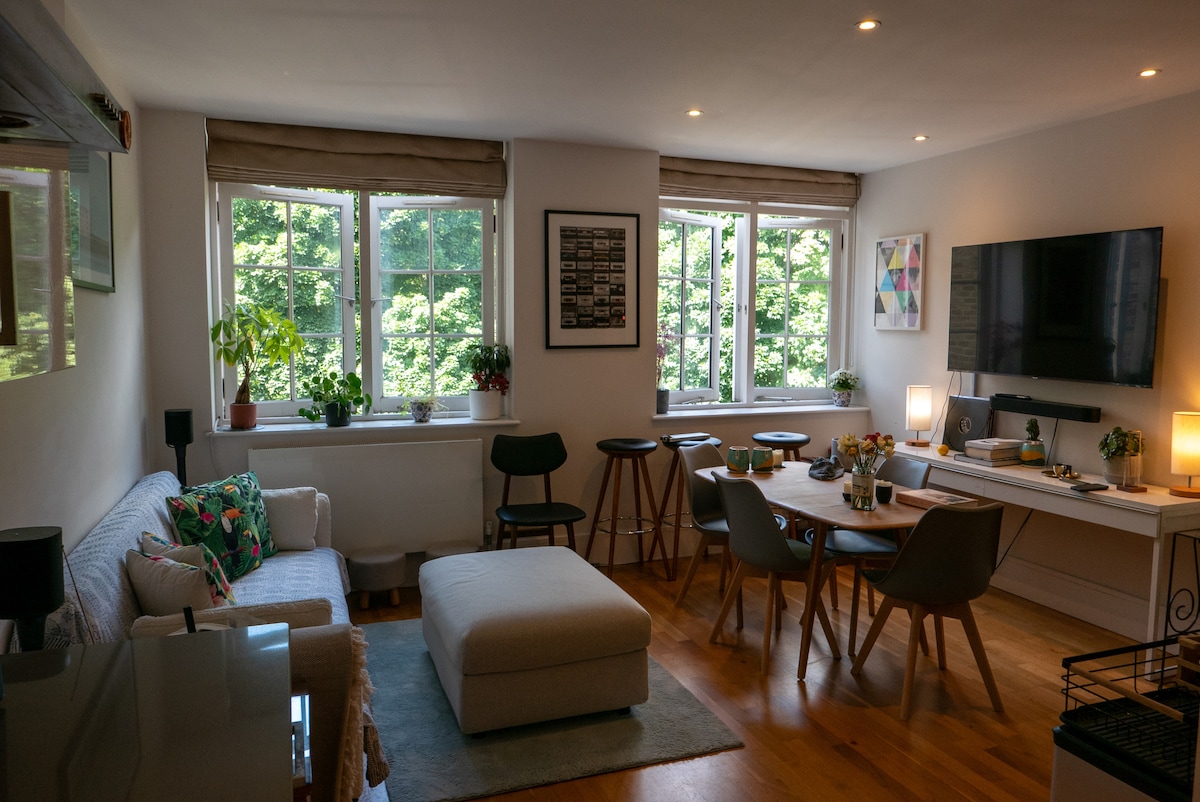 A comfortable living area features a light-colored sofa adorned with decorative pillows. Multiple windows provide natural light and a view of greenery. A wooden dining table is surrounded by chairs, while a wall-mounted TV and various plants add to the inviting ambiance.