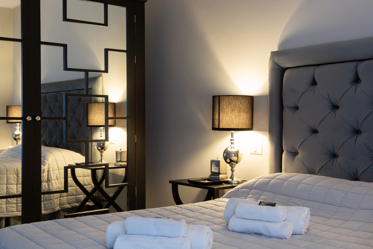 A spacious bedroom is presented with a comfortable bed adorned with neatly arranged towels. A mirrored wardrobe reflects soft lighting from stylish bedside lamps, creating a sophisticated ambiance. The elegant decor includes a muted color palette, enhancing the serene atmosphere of the space.