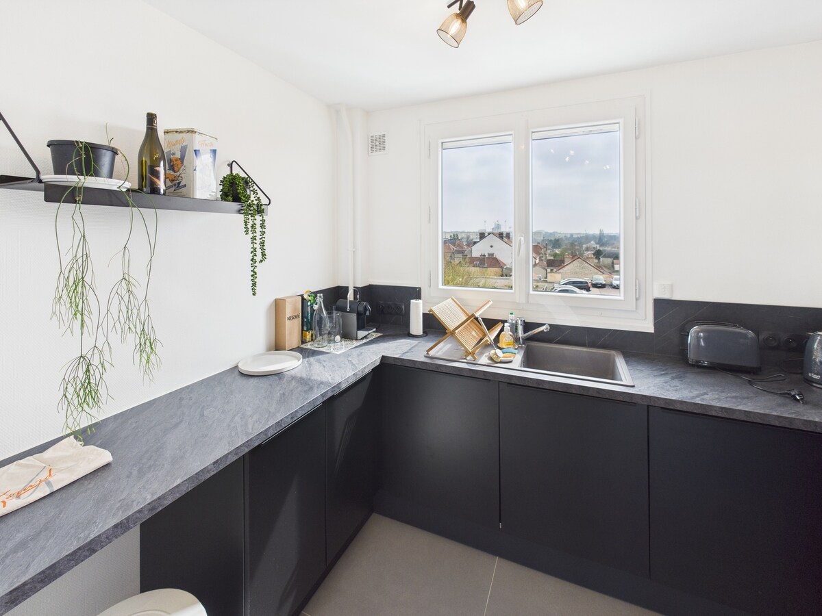 A modern kitchen featuring sleek black cabinets with a dark countertop. Natural light enters through a large window, highlighting the spacious layout. Shelves display minimal decor and plants. The area is equipped with essential appliances, ensuring a functional cooking space.
