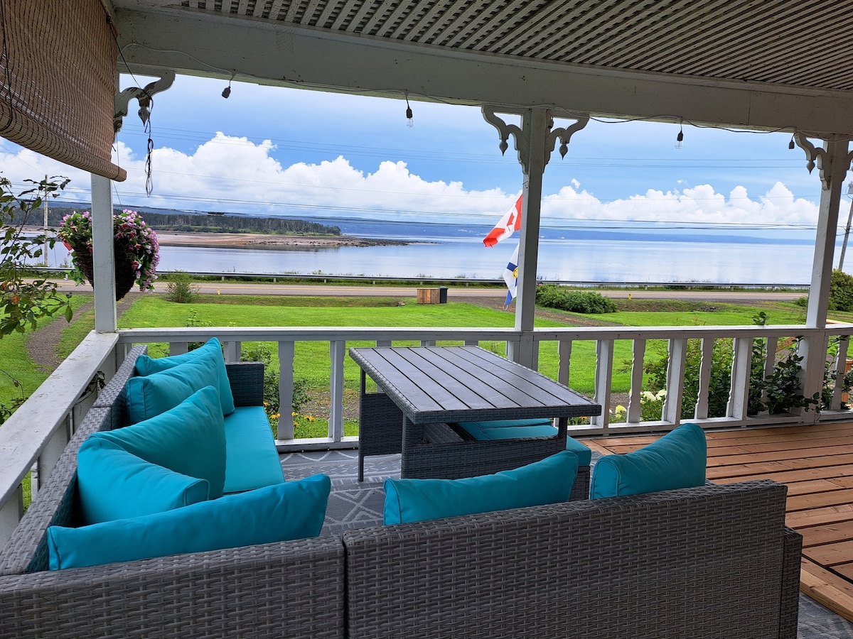 A spacious porch overlooks the tranquil Annapolis Basin, featuring a comfortable seating area with turquoise cushions. A wooden table is positioned in front, inviting guests to relax while enjoying the serene water views. The surrounding greenery and flowers enhance the peaceful ambiance.