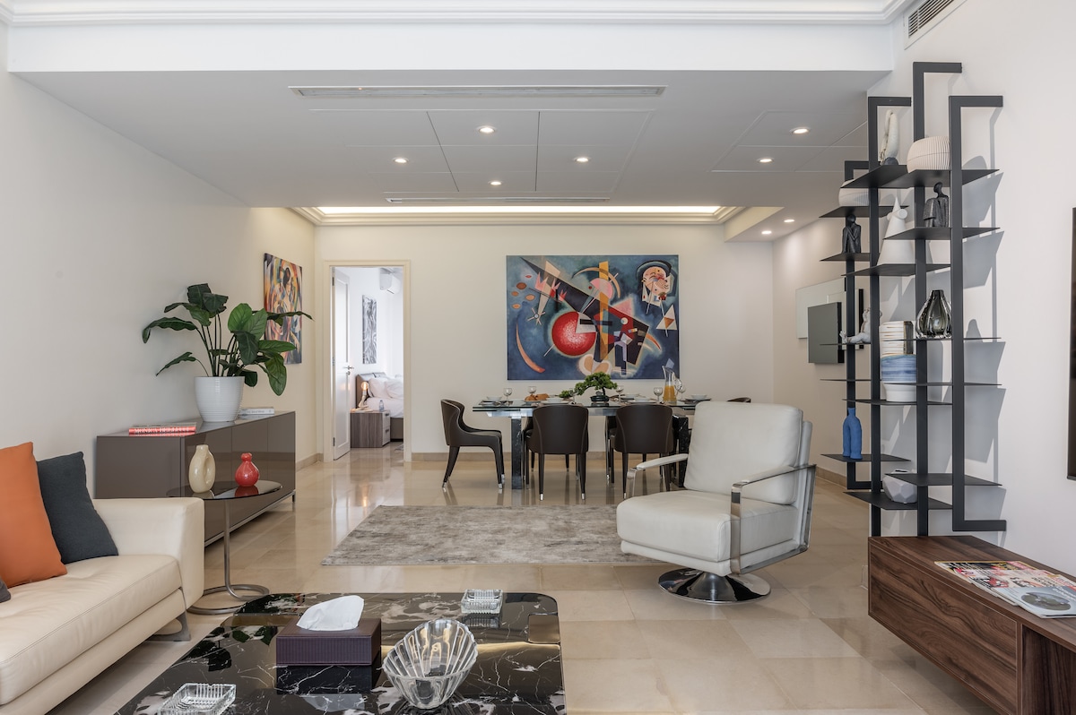An open-plan living and dining area is presented, featuring a modern beige sofa with colorful cushions. A large abstract artwork adorns the wall, while a dining table with stylish chairs is positioned nearby. A decorative shelf holds various artistic pieces, contributing to a contemporary aesthetic.