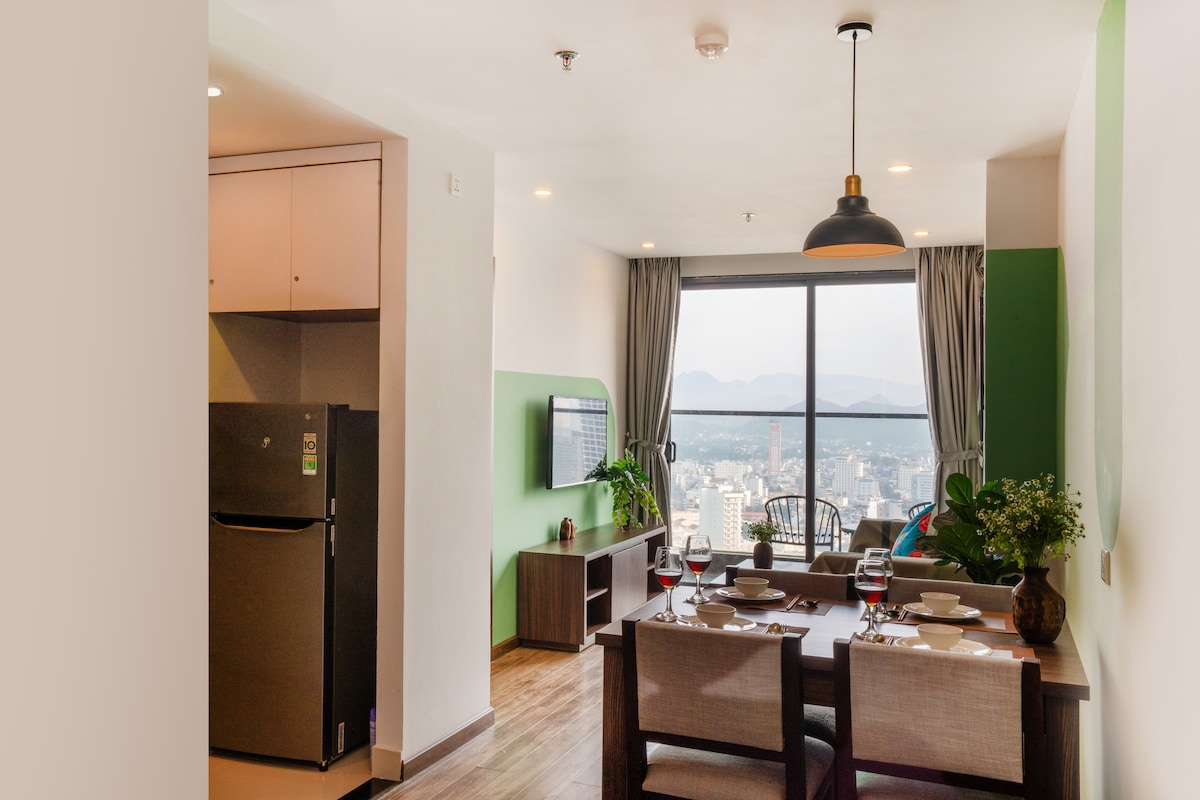 A modern living space displays a dining area with a wooden table set for four. A cozy lounge corner features a flat-screen TV and a well-equipped kitchen area. Large windows frame panoramic views of the city and mountains, providing ample natural light.