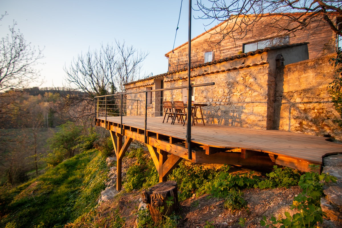 A wooden terrace is presented, elevated above lush greenery and surrounded by trees. The sun casts a warm glow on the weathered stone wall of the building behind. A simple wooden table and chairs are arranged on the terrace, offering a peaceful outdoor space.
