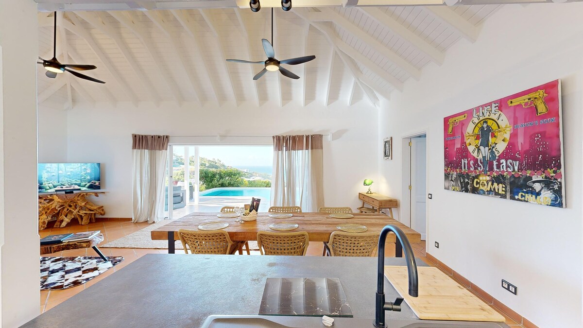 An open-concept kitchen and dining area is presented, featuring a long wooden dining table surrounded by wicker chairs. Bright natural light fills the space, highlighting the vaulted ceiling and ceiling fans. Views of the swimming pool and garden are visible through large sliding doors.