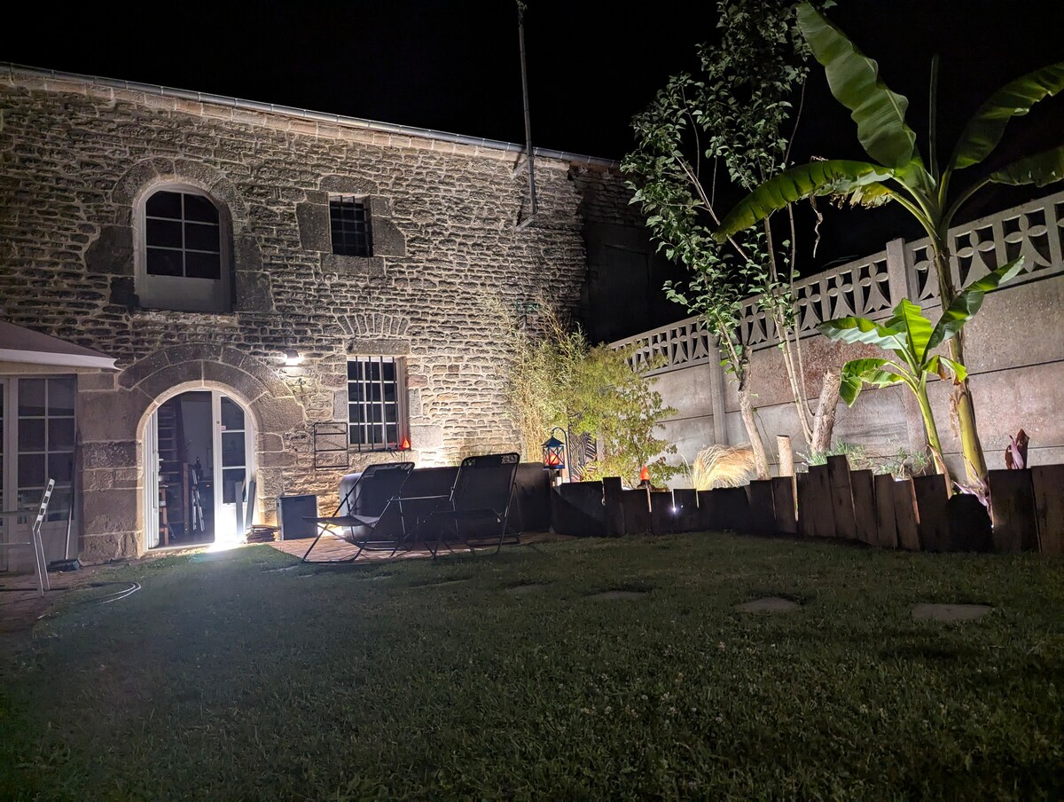 The image shows a charming house with exposed stone walls, illuminated by soft outdoor lighting. A serene garden space features multiple lounge chairs arranged for relaxation. Lush greenery, including banana plants, can be seen alongside a wooden fence, contributing to a peaceful outdoor setting.