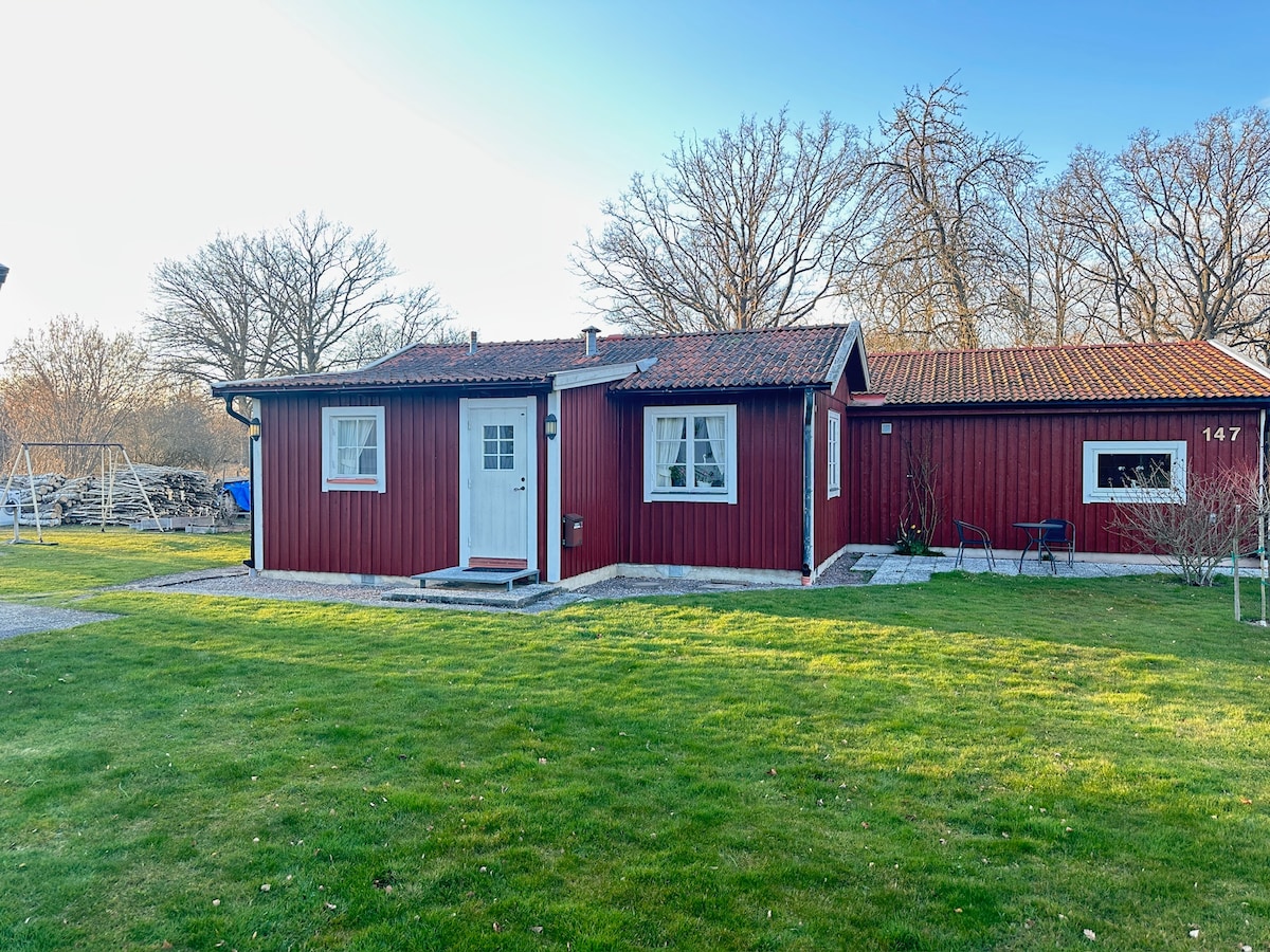 The red cottage, featuring a white door and framed windows, sits within a spacious green lawn. A pathway leads to the entrance, and nearby, a swing set can be seen in a natural setting with trees in the background.