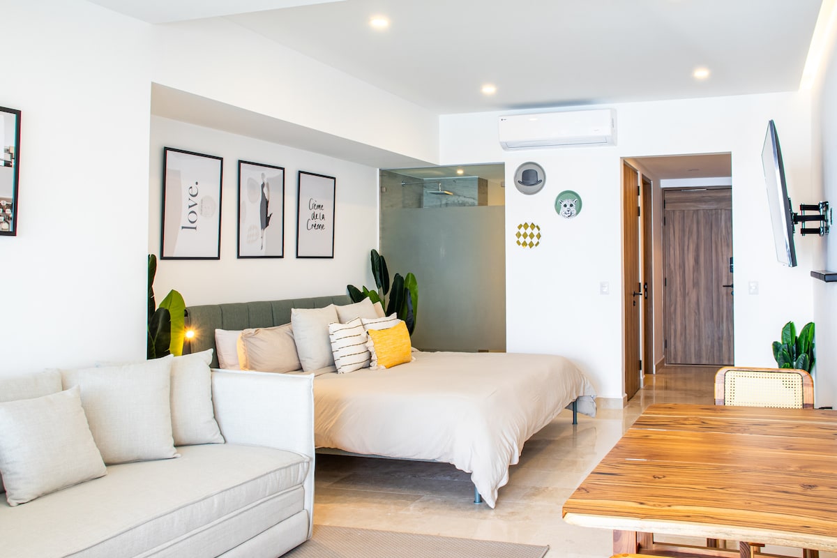 A bright living area features a spacious bed with accent pillows and a minimalist design. A comfortable sofa is positioned nearby, complemented by a wooden dining table. Wall art and plants enhance the aesthetic, while soft lighting creates an inviting atmosphere.