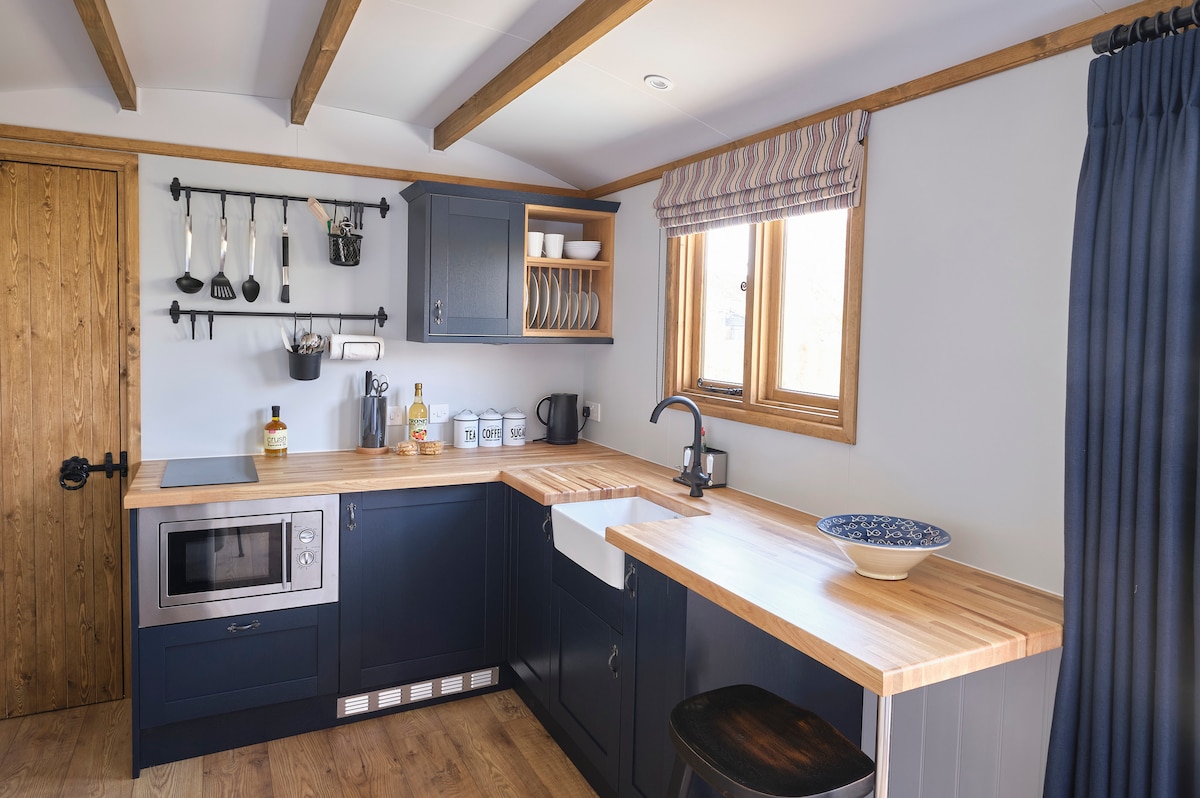 A well-equipped kitchen features wooden countertops and navy blue cabinetry. Essential appliances include a microwave and sink. Cookware is organized on wall-mounted hooks. Natural light enters through a window adorned with striped fabric. A cozy dining area is present with a bowl on the countertop.