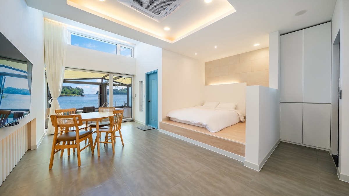 A spacious room features a raised sleeping area with a light-colored bedding arrangement. A dining area with wooden chairs and a round table is positioned nearby. The large windows offer views of the water, allowing natural light to fill the space.