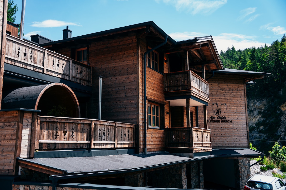 A multi-level wooden structure is featured, showcasing balconies with wooden railings. The building's rustic design is complemented by surrounding greenery and a clear blue sky. Various textures of wood are visible, enhancing the natural aesthetic of the property.
