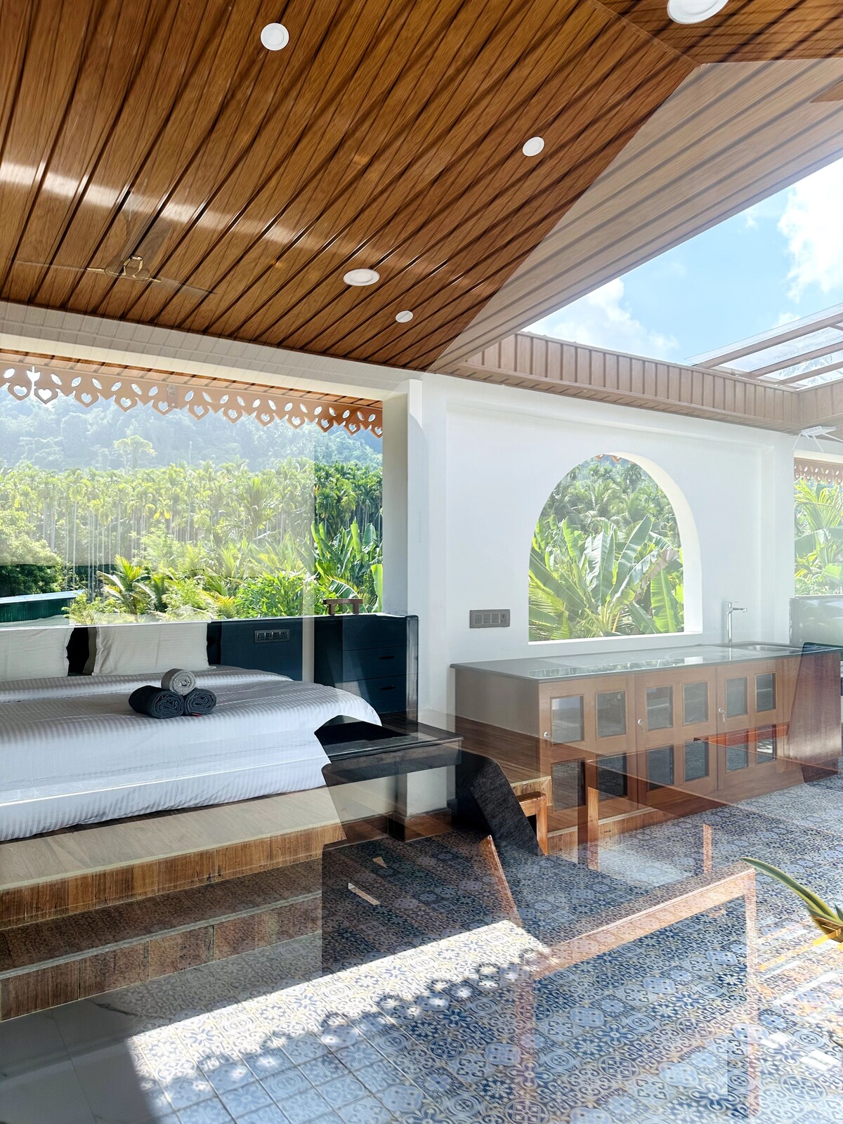 A spacious area is highlighted by a large bed with neatly arranged linens and decorative towels. Glass doors open to views of lush greenery, allowing natural light to fill the room. A modern kitchenette is visible, complemented by decorative tile flooring.
