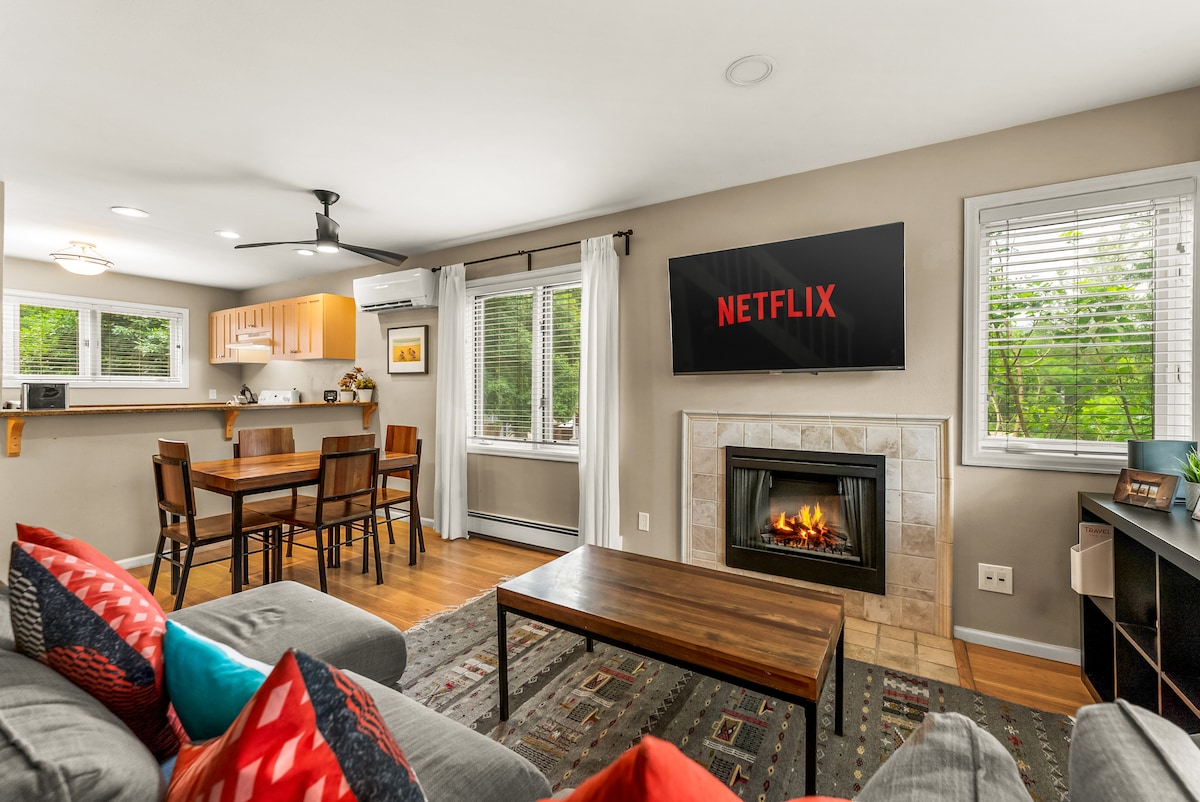 The living area features a comfortable sofa with colorful cushions and a modern design. A wall-mounted television displays Netflix, while a dining table for four is positioned nearby. Natural light enters through two windows, creating a warm ambiance, complemented by a stylish fireplace.