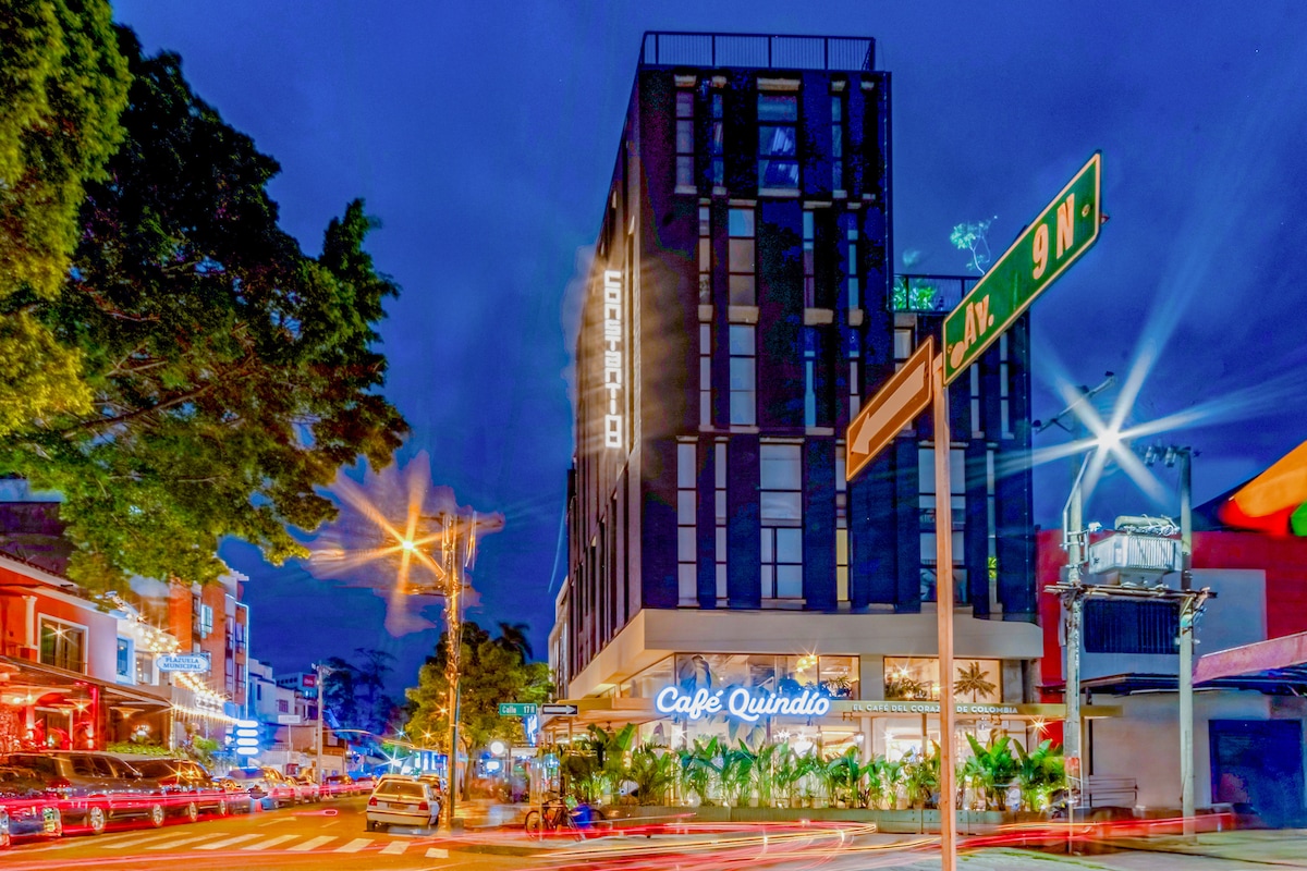 The modern exterior of the Constantino building is presented at night, illuminated by soft lighting. The Café Quindío sign is prominently displayed, and street signage indicates proximity to nearby attractions. Lush greenery decorates the entrance, contributing to the vibrant atmosphere of the surrounding area.