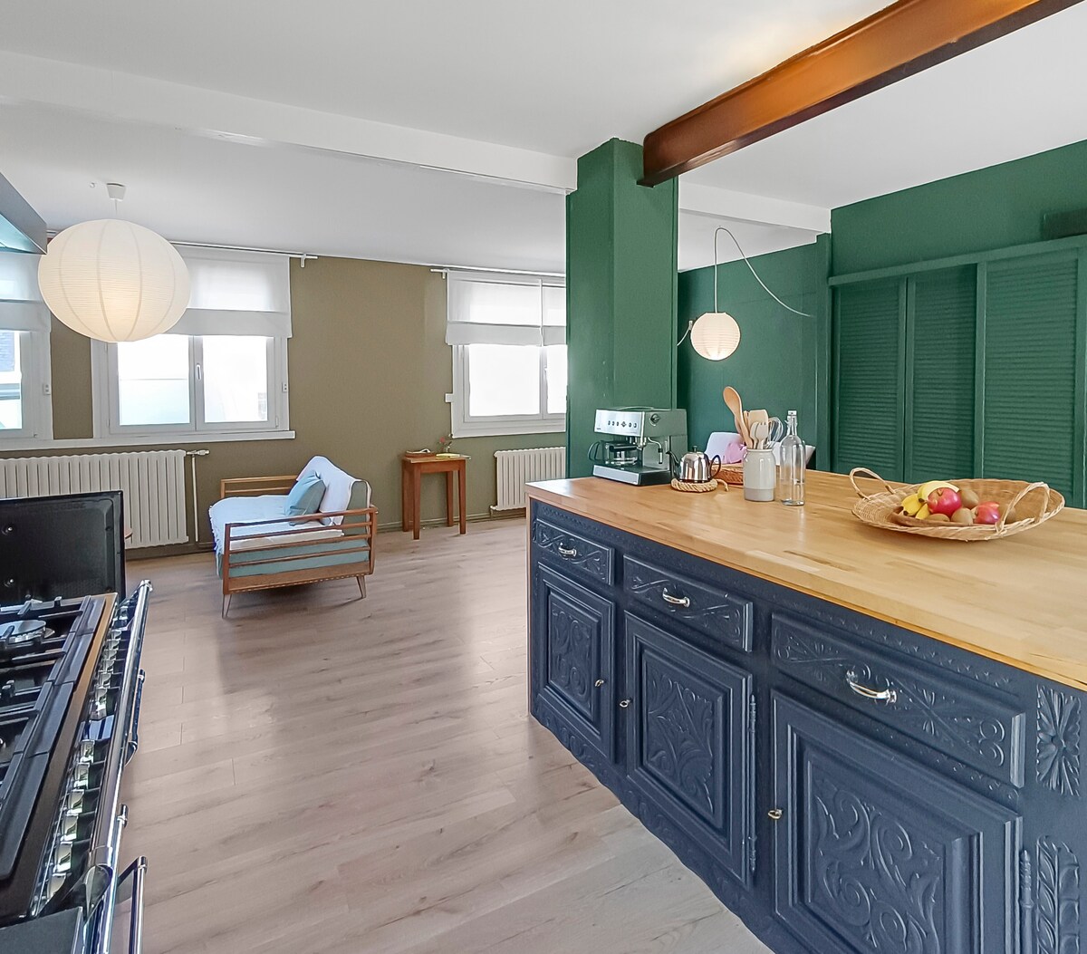 A spacious kitchen area is depicted, featuring a large central island with a wooden countertop. Dark cabinetry contrasts with light walls. A cozy seating area is present in the background, complemented by a wooden table. Natural light filters through the windows, enhancing the room's atmosphere.