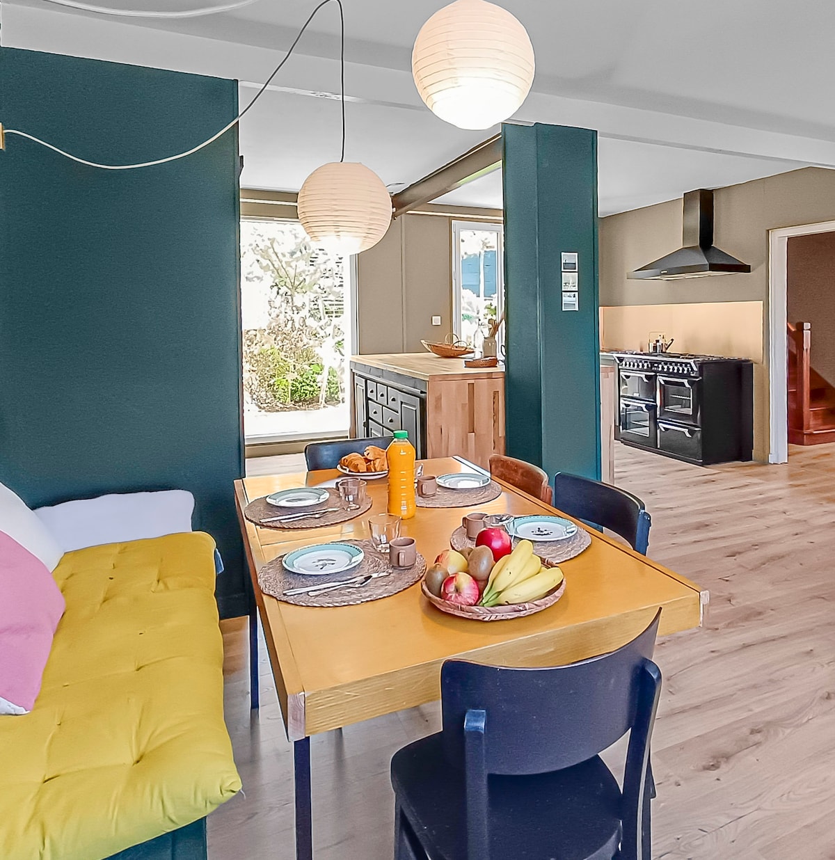 The kitchen area displays a wooden dining table set for four, featuring plates and a bowl of fresh fruit. A cozy yellow sofa is positioned nearby, with large windows allowing natural light to enhance the space. The kitchen is visible in the background, showcasing an equipped cooking area.