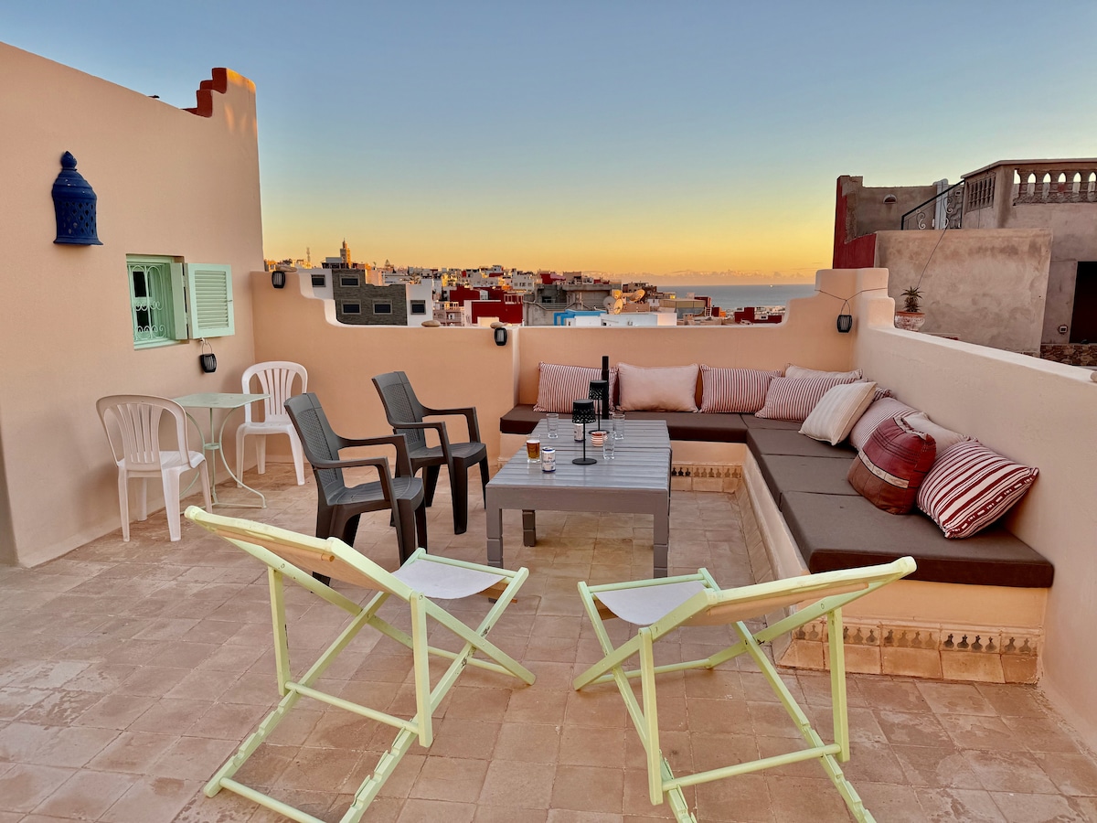 A rooftop terrace offers a relaxing seating area with a combination of lounge cushions and chairs, accompanied by a low wooden table. The view includes distant buildings and the sea, illuminated by a soft sunset glow.