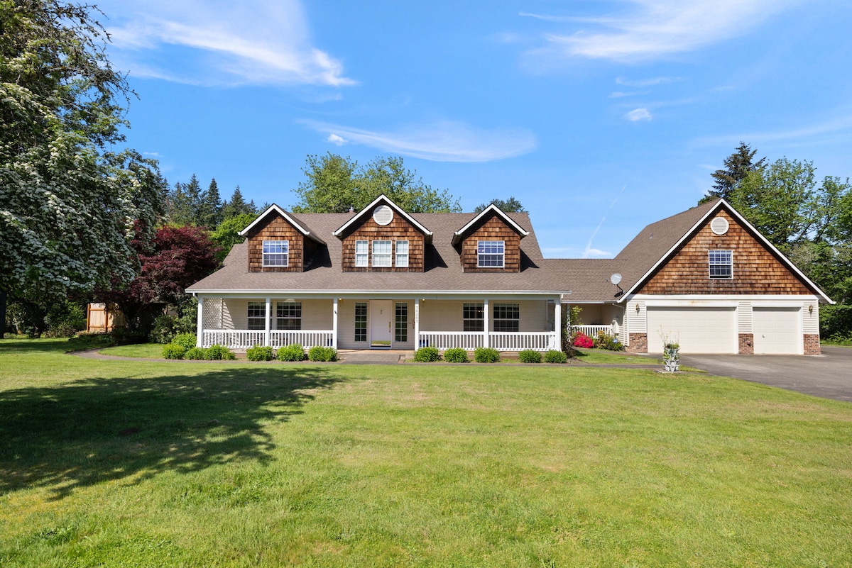 The charming farmhouse features a welcoming front porch with railing, surrounded by a spacious lawn. The two-story structure is complemented by a mixture of wood siding and shingles, along with multiple windows that enhance natural light. A paved driveway leads to a two-car garage.