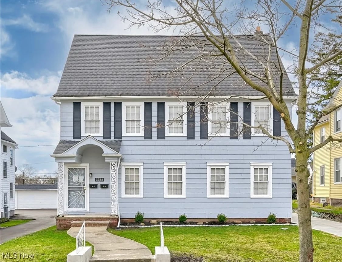 A two-story blue house features a classic design with white trim and five large, evenly spaced windows on the first and second floors. A well-maintained lawn is visible in front, along with a concrete pathway leading to the entrance.