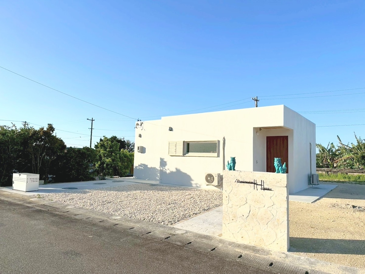 A modern white house is prominently positioned on a gravel lot. The building features a distinctive entrance with wooden doors and a large window allowing natural light. Surrounding greenery adds a touch of nature, and clear blue skies create a bright atmosphere.