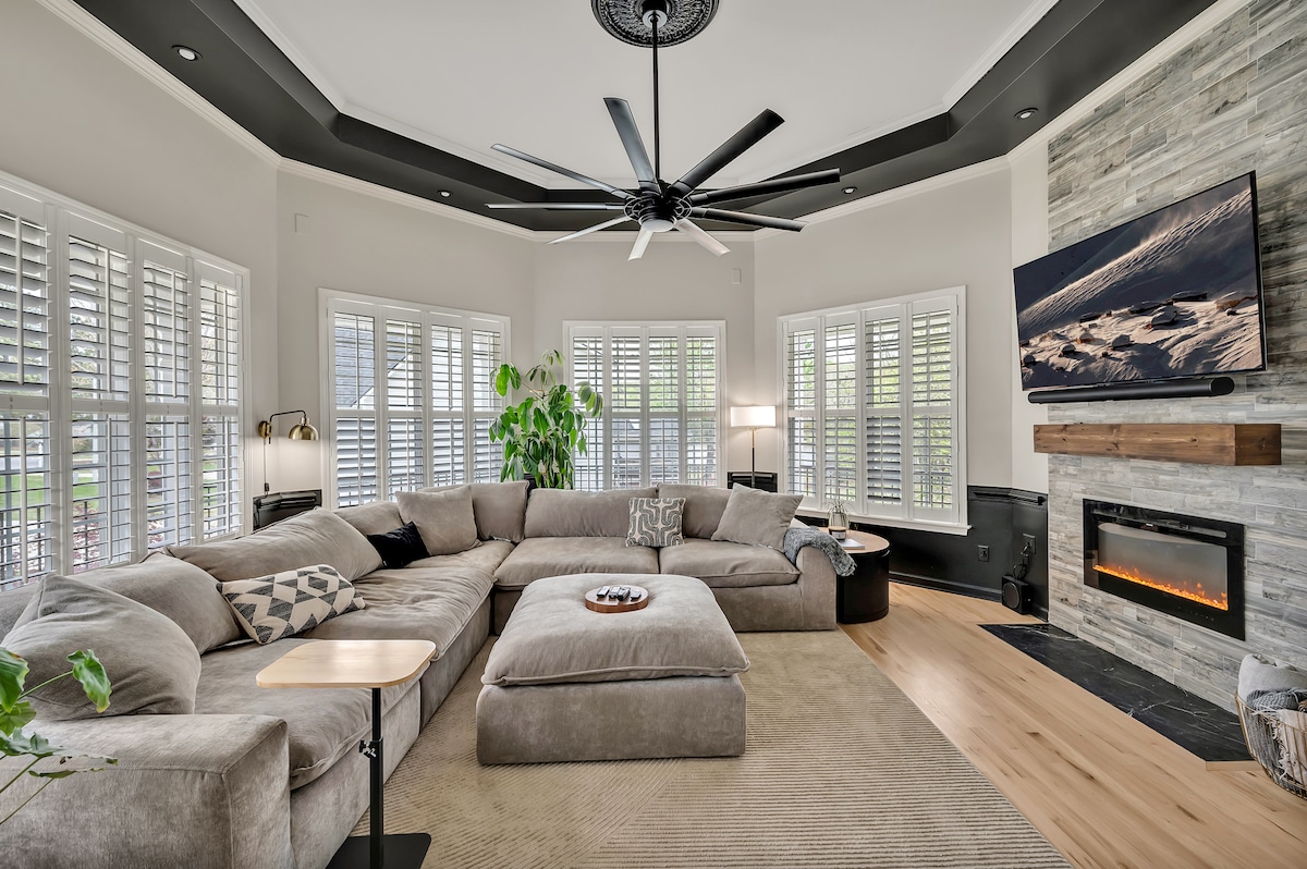 A spacious living room features a large sectional sofa, a contemporary ceiling fan, and a flat-screen TV mounted above a modern fireplace. Natural light enters through multiple windows adorned with shutters, while a potted plant adds greenery to the inviting atmosphere.