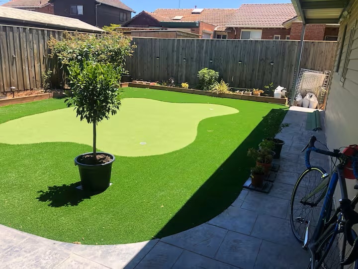 A private outdoor space features a vibrant green artificial turf area, complemented by a small potted tree at its center. Planters with various plants can be seen along the edges. The area is bordered by a wooden fence and a tiled pathway, with a bicycle resting nearby.
