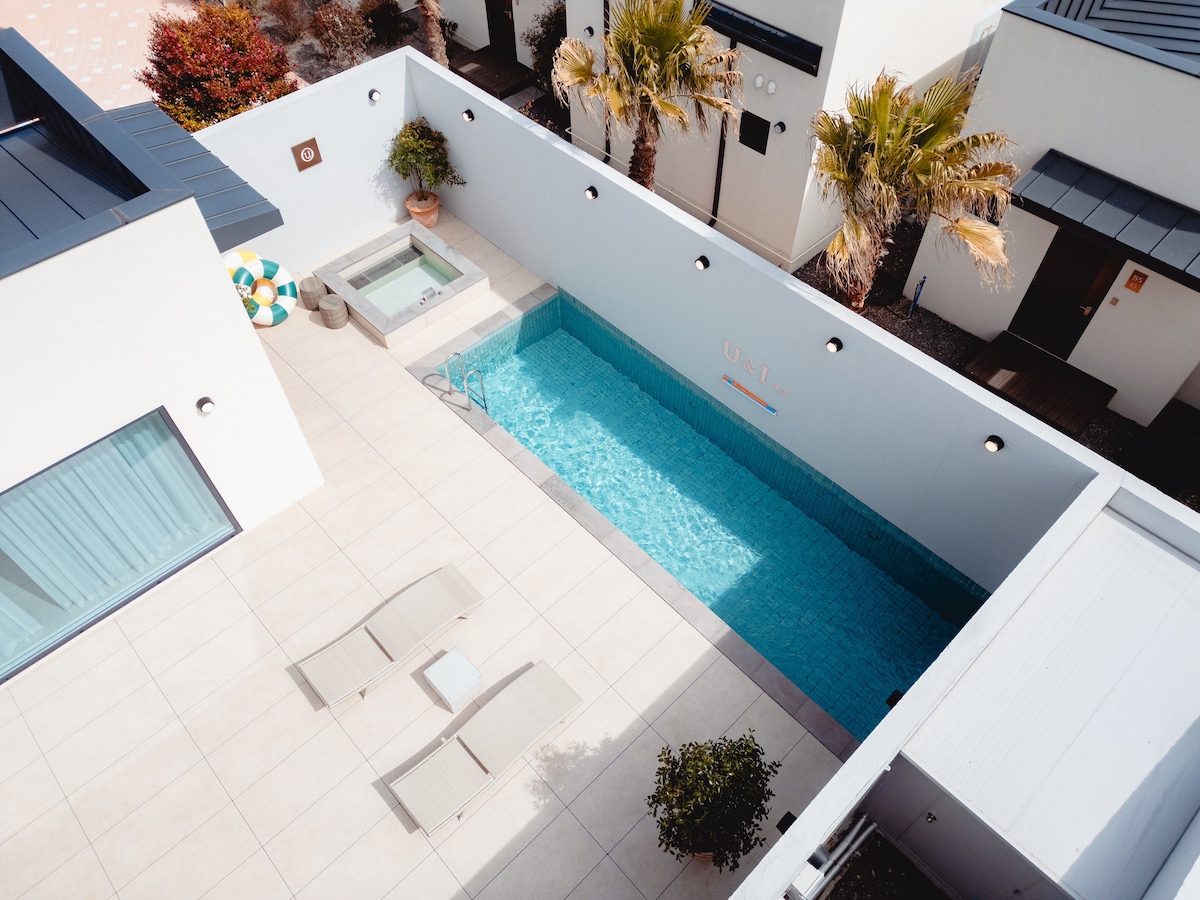 An aerial view displays a serene outdoor area featuring a private swimming pool bordered by sun loungers. A small hot tub adds to the relaxation space. Lush greenery and palm trees provide a tropical ambiance, while the clean, paved surface enhances the overall tranquility of the setting.