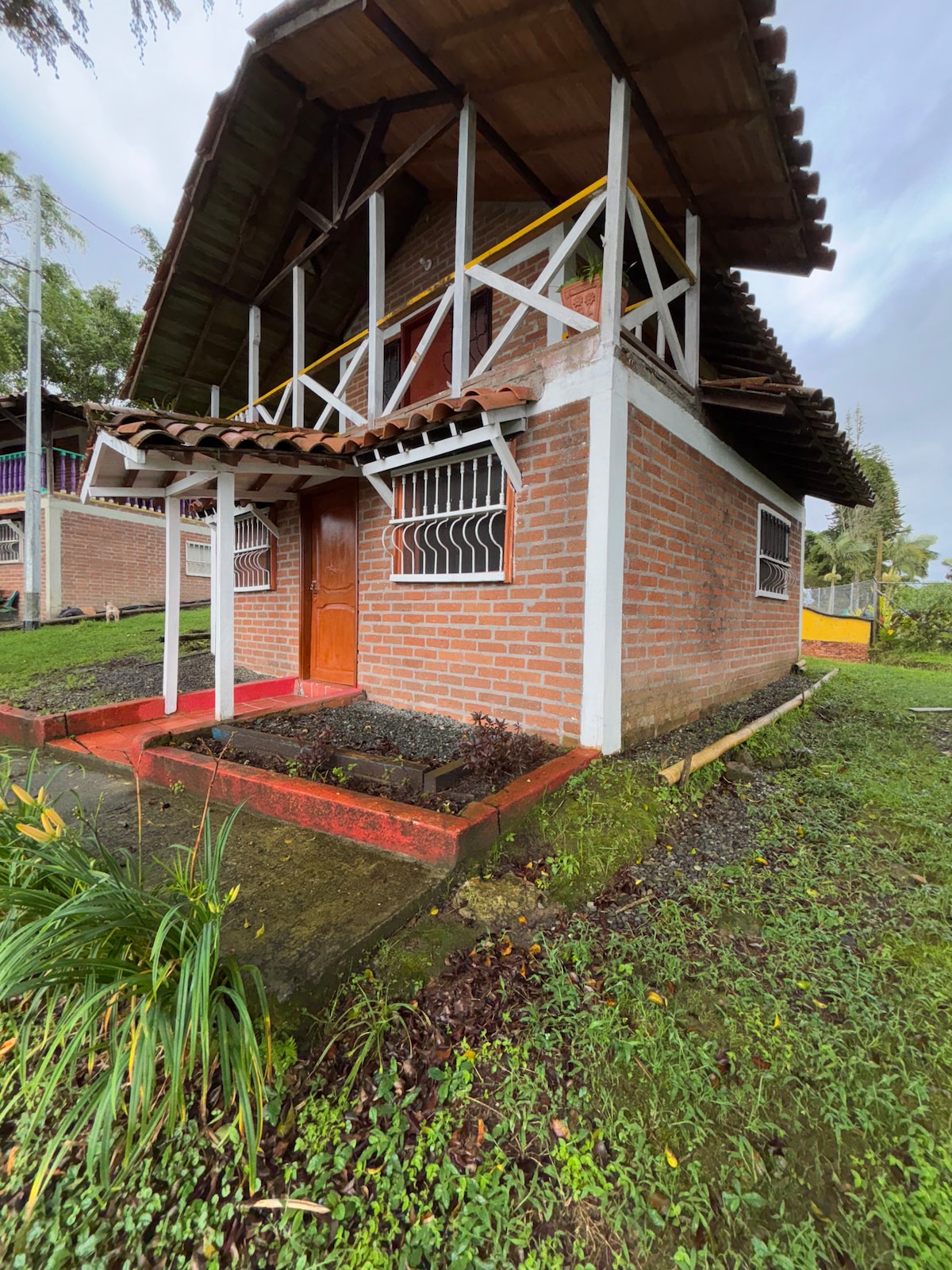 A two-story cabin is showcased surrounded by green grass and natural vegetation. The structure features a brick facade with wooden accents. A welcoming wooden door is positioned at the entrance, under a roof with overhanging eaves, enhancing the rustic charm of the setting.