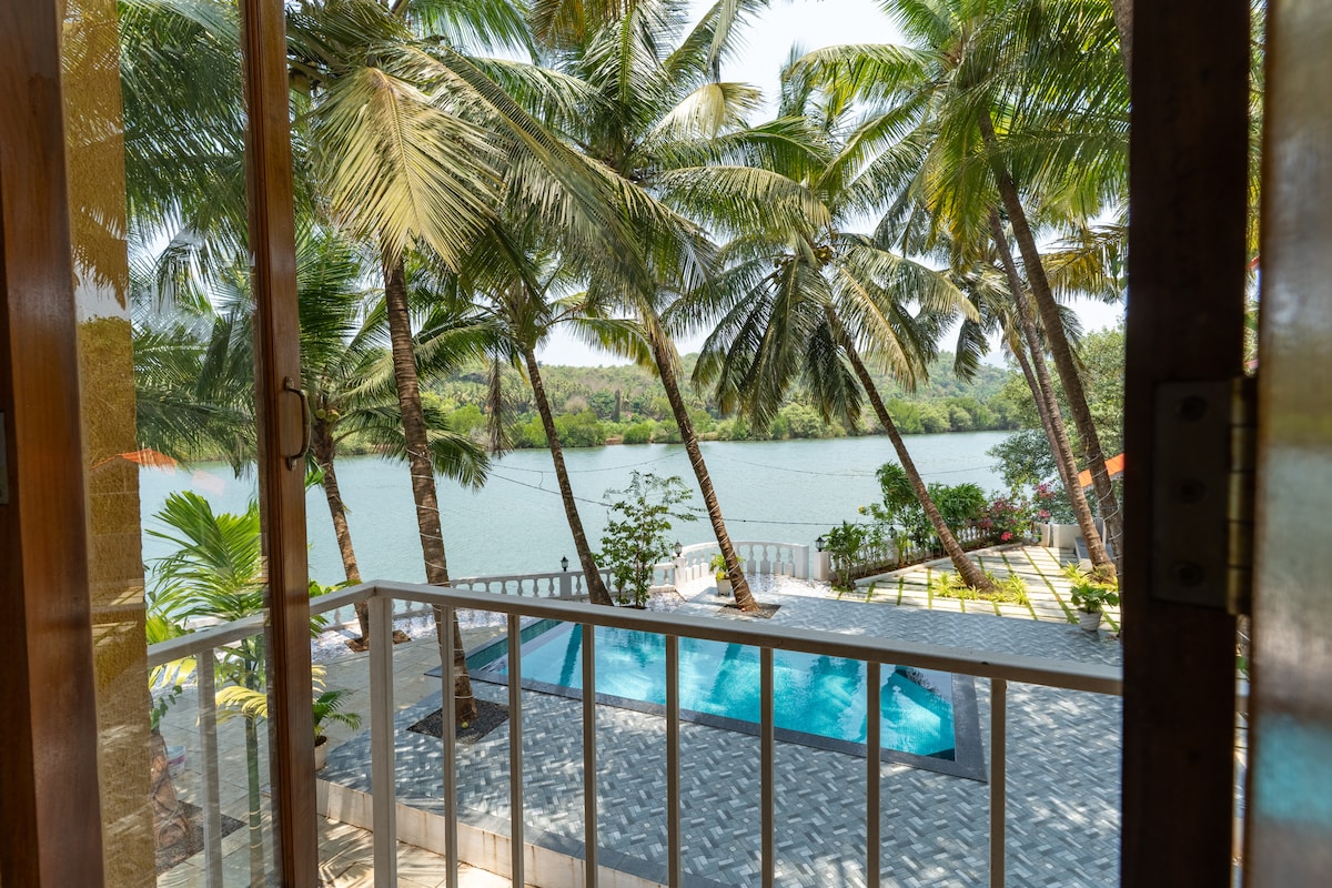 A view from the private riverside balcony showcases swaying coconut trees framing the serene Talpona River. The sparkling swimming pool is visible below, complemented by lush greenery and a sunlit pathway leading to the water, creating a peaceful outdoor atmosphere.