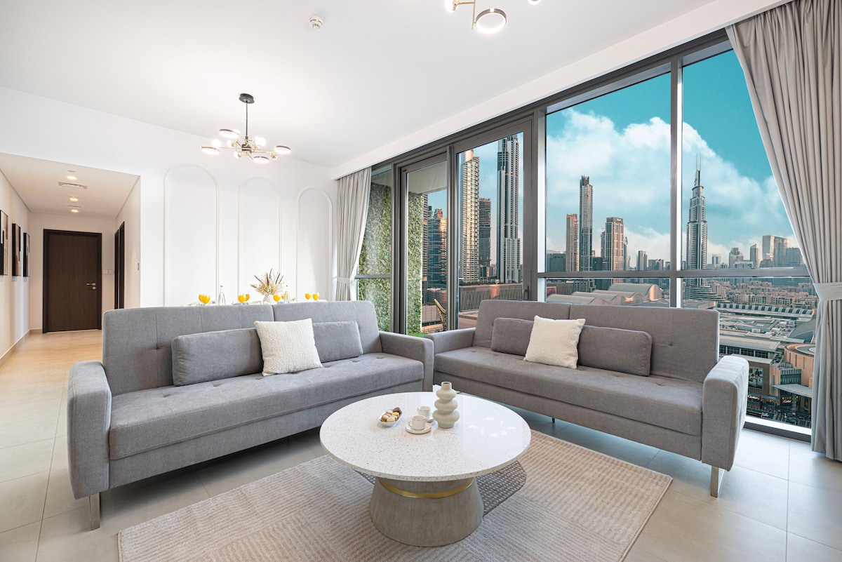 A spacious living room features two gray convertible sofa beds and a round marble coffee table. Full-height windows provide uninterrupted views of the Burj Khalifa and the skyline, while elegant curtains frame the scene. Natural light brightens the modern decor.