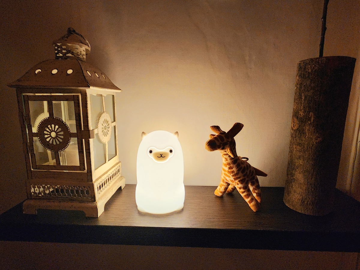 A small shelf displays a soft, glowing alpaca lamp in the center, flanked by a decorative lantern on the left and a plush giraffe toy on the right. The warm light creates a gentle ambiance against the dark backdrop.