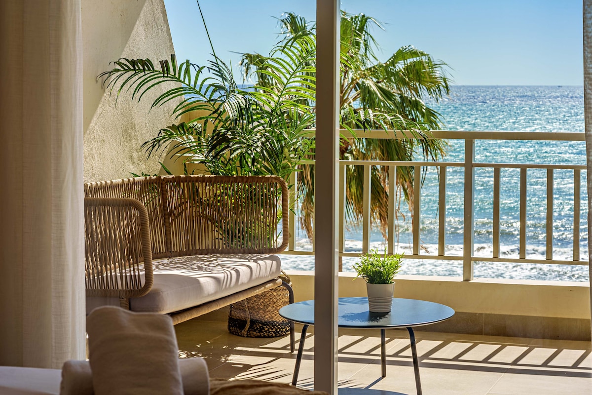 A cozy seating area features a rattan bench and a round coffee table. Lush greenery is observed in the background, complemented by the ocean waves visible beyond the balcony railing. Bright sunlight illuminates the space, creating a serene atmosphere.