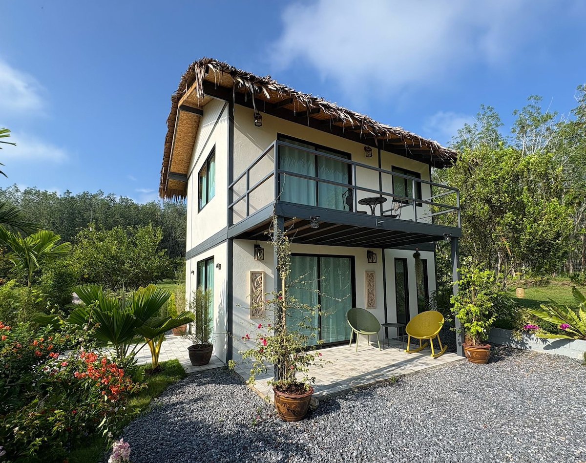 The image showcases a two-story building featuring a thatched roof and large glass doors on the ground level. A balcony extends from the upper floor, complemented by two chairs. Surrounding gardens filled with tropical plants and vibrant flowers enhance the natural setting.