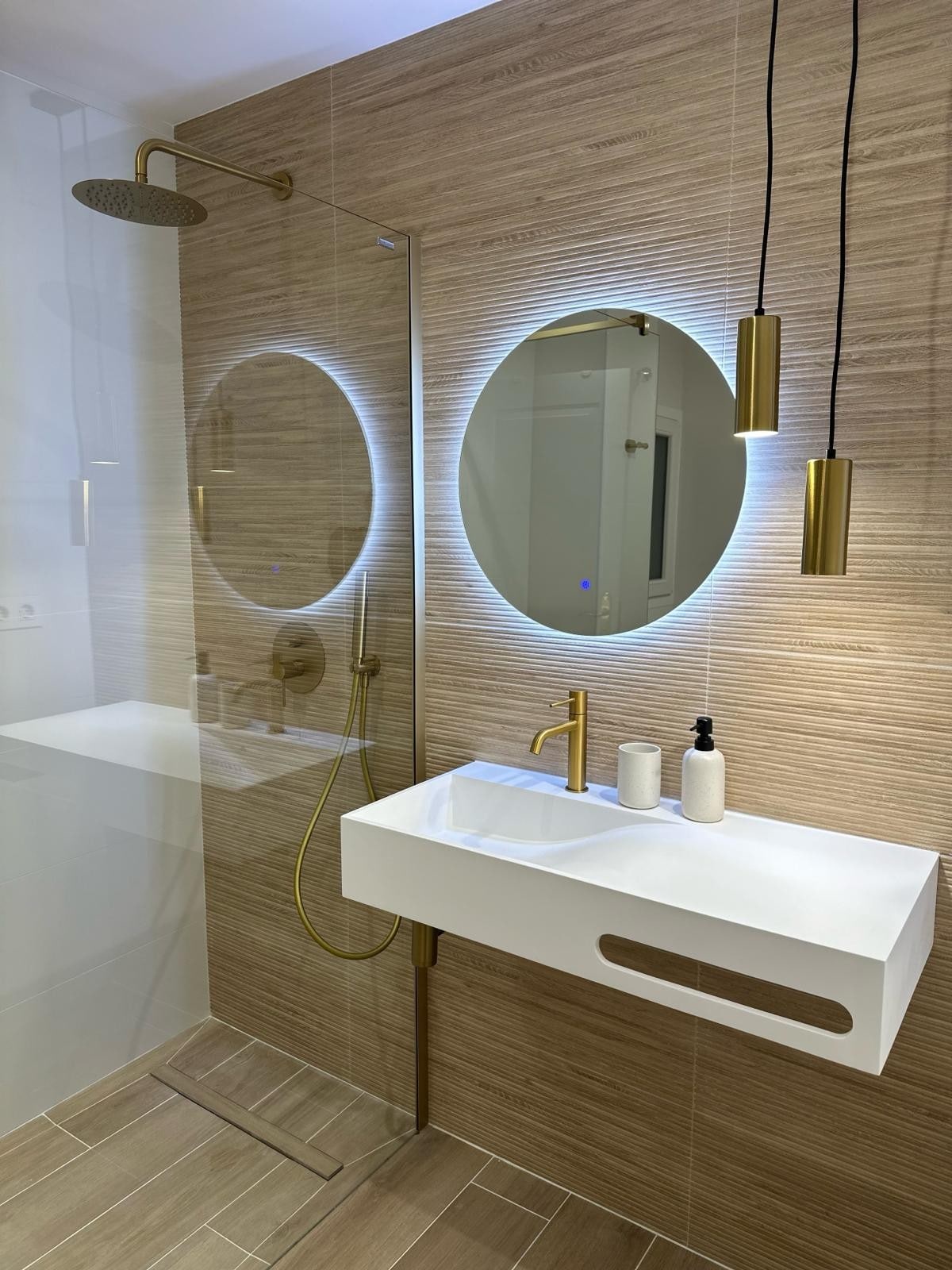 The modern bathroom features a sleek, wall-mounted sink with a minimalist design and gold accents. A circular mirror is illuminated by soft light, complementing the elegant shower area with a rain shower head. Neutral-toned tiles add warmth to the inviting space.
