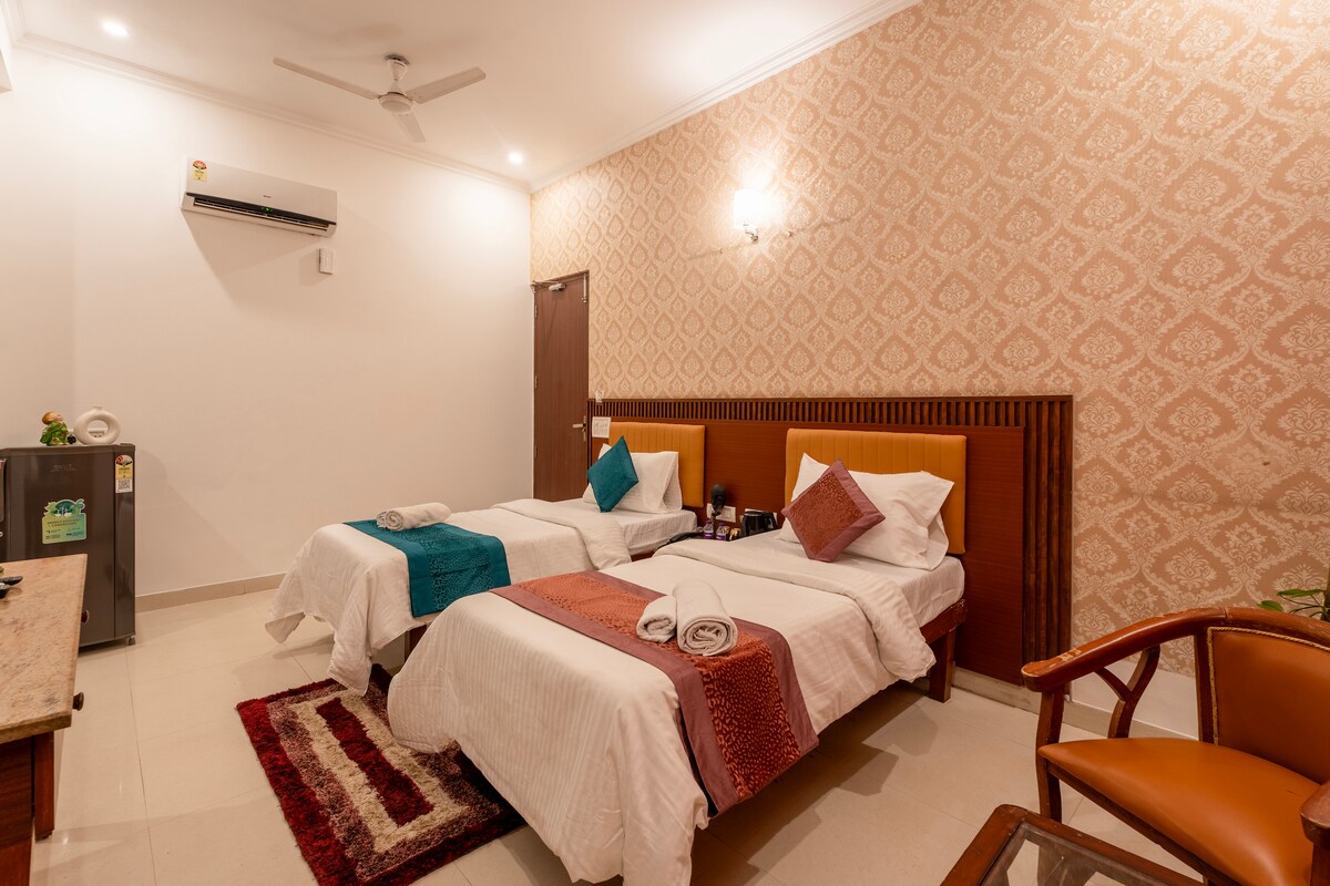 The studio apartment features two spacious beds dressed in white linens, complemented by decorative throws in teal and terracotta. A small refrigerator is present beside the door, with a wooden chair positioned next to a subtle area rug, adding comfort and functionality to the space.