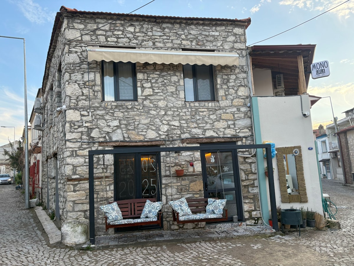 A charming stone building features a welcoming façade, complemented by a covered entrance. Two wooden chairs with decorative cushions are positioned in front, inviting relaxation. The surrounding cobblestone street and the building's rustic texture contribute to the neighborhood's character.