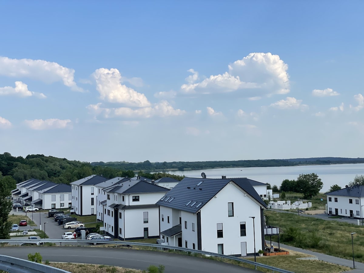 A view from a hill showcases modern white buildings arranged in clusters, set against a backdrop of a calm lake. Lush green landscapes frame the scene, while fluffy clouds drift in a blue sky above the tranquil waters.